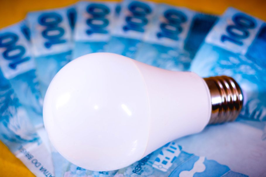 Lâmpada LED branca sobre várias cédulas de 100 reais dispostas em leque, simbolizando o aumento no custo da energia elétrica.
