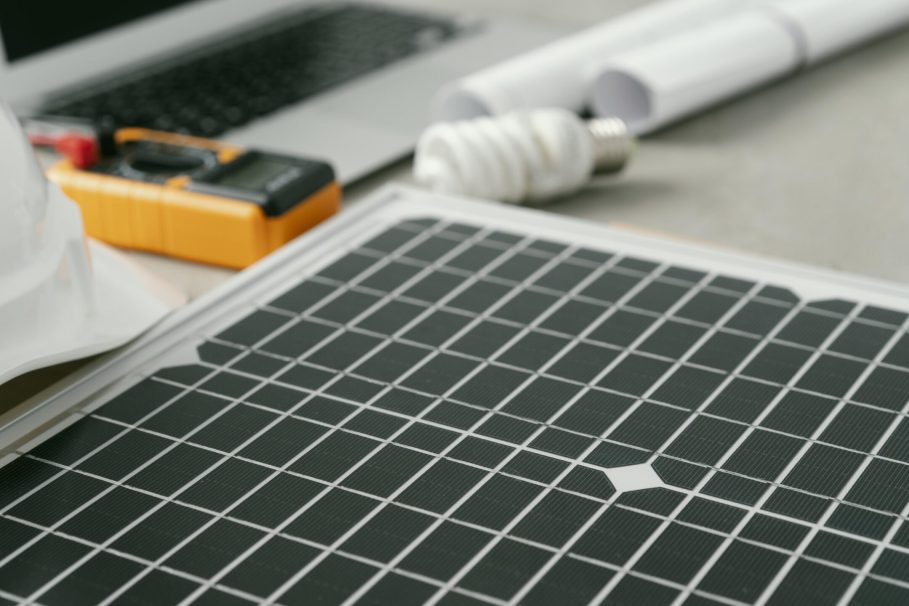 high-angle-eco-plan-arrangement Equipamentos de energia solar sobre uma mesa, incluindo um pequeno painel solar, capacete de segurança, lâmpada, multímetro, notebook e projetos técnicos enrolados.