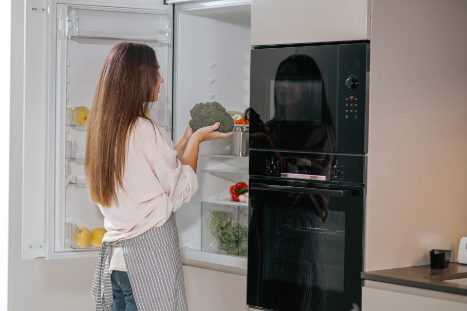 Young housewife standing near the fridge in the kitchen Mulher de costas, usando avental listrado, segura um brócolis na frente de uma geladeira aberta em uma cozinha moderna.