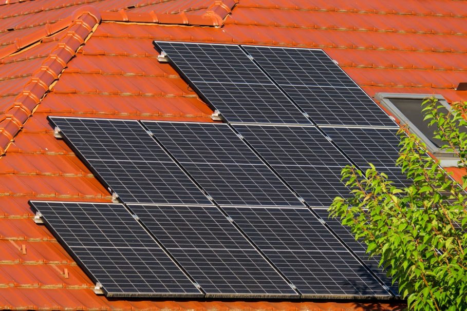 Painéis solares instalados em telhado de telhas cerâmicas alaranjadas com vegetação ao lado.