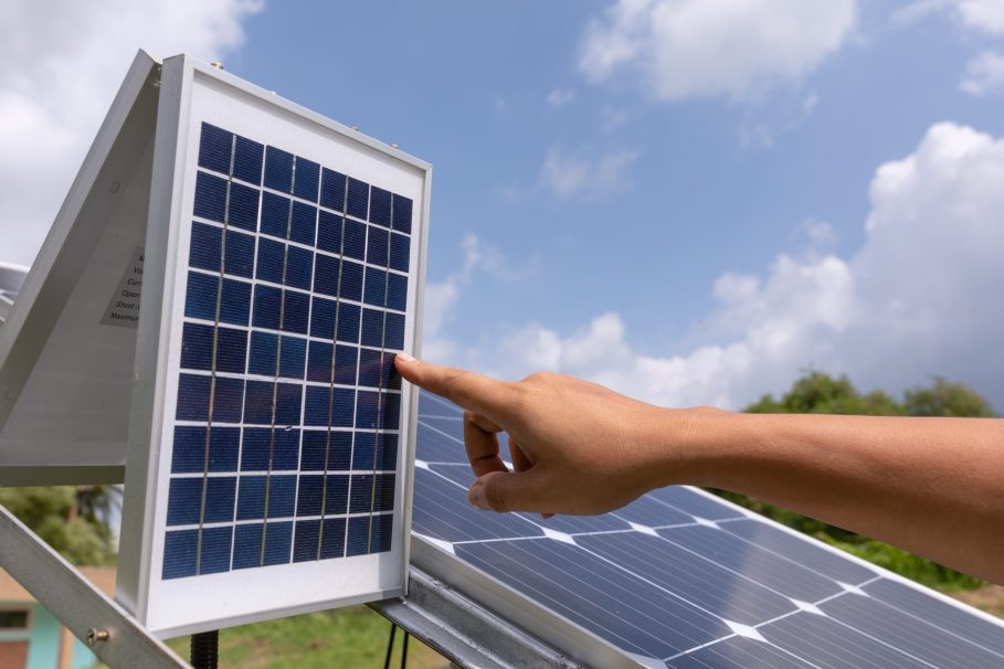 Mão apontando para um painel solar fotovoltaico em um ambiente ao ar livre, com céu azul, nuvens leves e vegetação ao fundo.