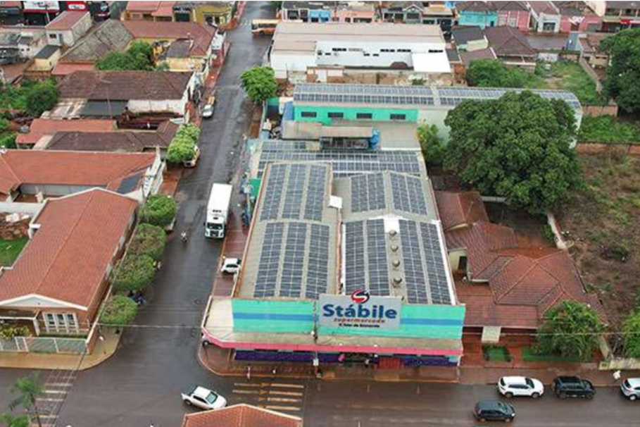 Vista aérea de um supermercado chamado "Stábile" com placas solares no telhado, cercado por casas e árvores em uma área urbana.